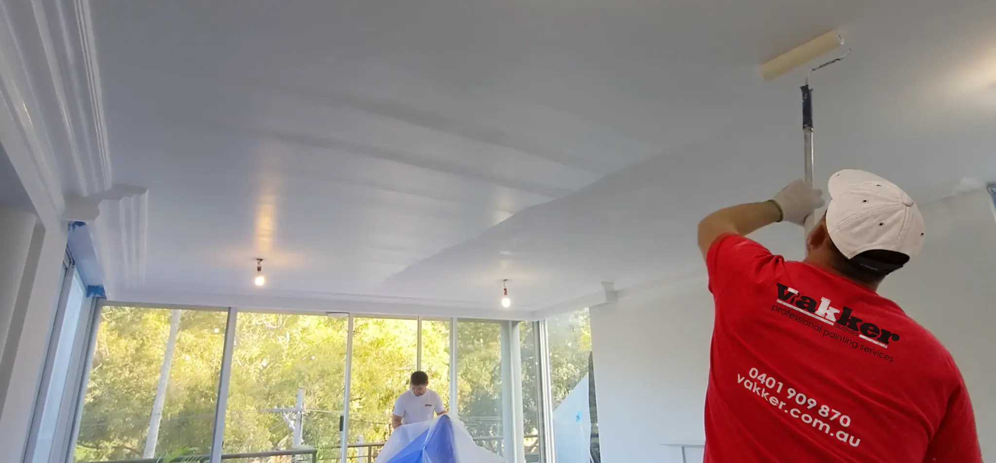 Vakker painter rolling a ceiling while a second painter covers furniture with protective sheeting.