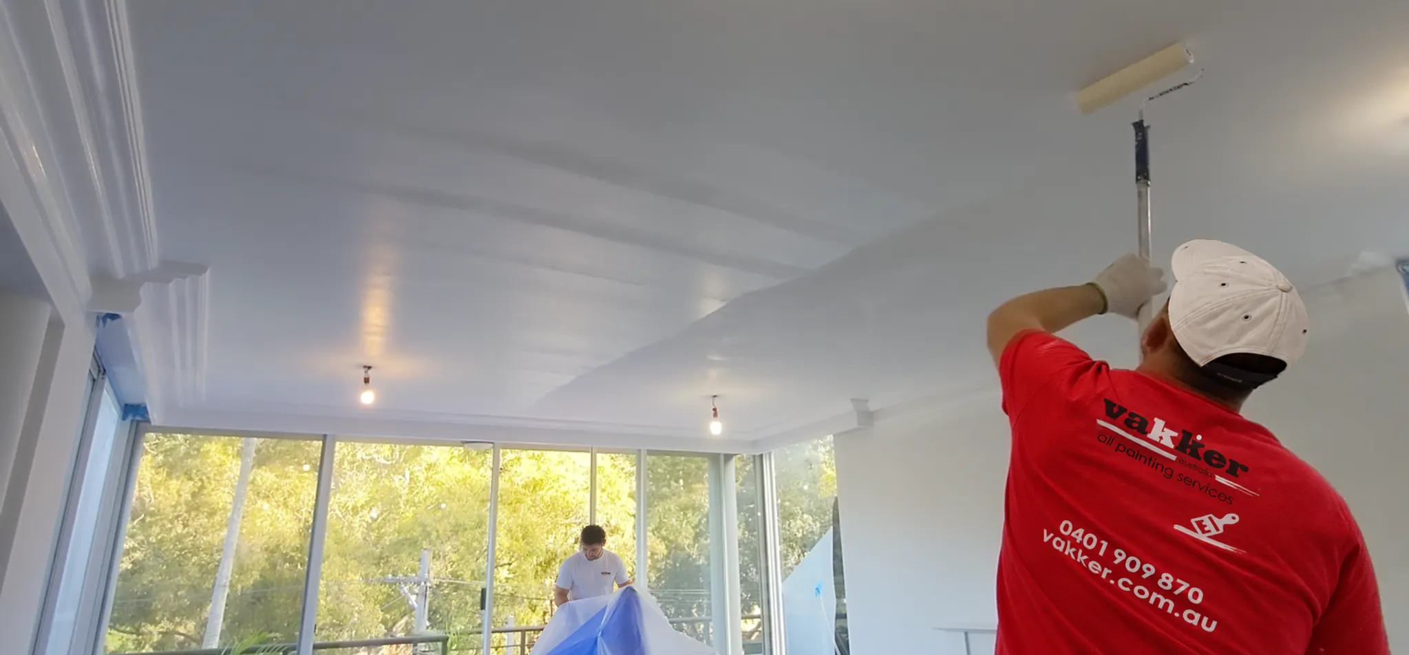 Vakker painter rolling a ceiling while a second painter covers furniture with protective sheeting.